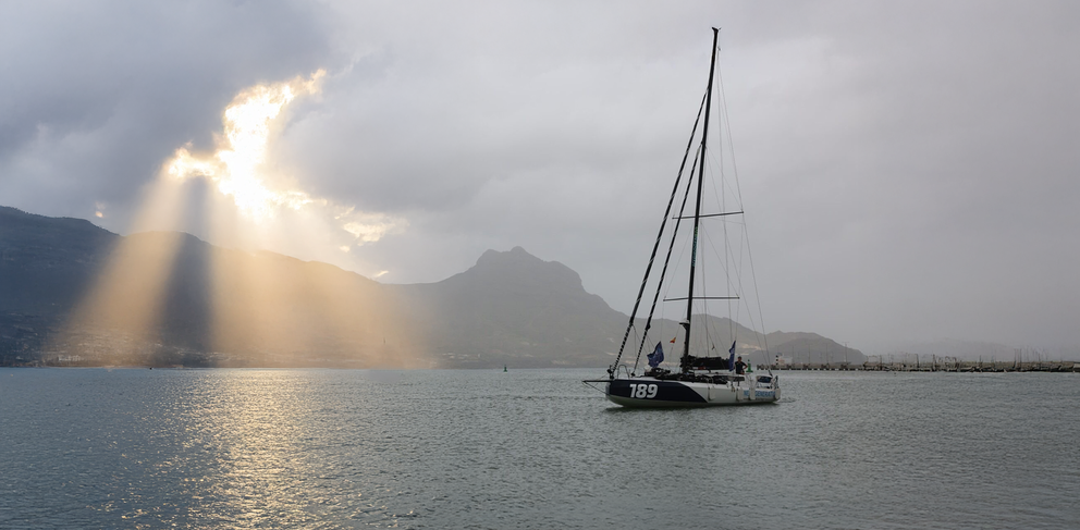 Ein Segelboot mit der Nummer 189 segelt auf einem ruhigen Gewässer, mit Bergen im Hintergrund und dramatischen Sonnenstrahlen, die durch die Wolken brechen und einen Teil der Landschaft beleuchten.